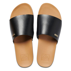 A pair of REEF Cushion Scout slides in brown and black, featuring a single wide black vegan leather strap over the foot. The tan footbed showcases the "Reef" brand logo embossed near the heel and includes a cushion bounce midsole for added comfort. Each strap is adorned with a small metallic "Reef" tag, photographed from above.