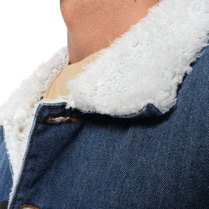 A close-up image of a person wearing the Ringers Western Duncan Denim Jacket, featuring the signature white, fluffy sherpa lining over a beige shirt. The focus is on the jacket's upper section and the texture of the lining. The person's face is not visible.