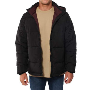 A person wearing a black Ringers Western Bairnsdale Puffer Jacket over a beige t-shirt and blue jeans stands facing forward. The jacket, which features a detachable hood and high collar for extra warmth, is unzipped to reveal the t-shirt underneath. The background is plain white.