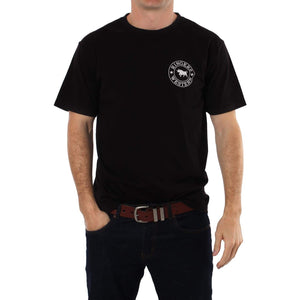 A man is standing and wearing a Ringers Western Signature Bull Classic T-Shirt, which is a comfortable black crew neck with a small white circular logo on the left side of the chest, featuring the text "Ringers Western" and an illustration of a bull. He has one hand in his pocket and is also wearing dark blue jeans and a brown belt.