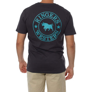 A person stands facing away, wearing the comfortable Ringers Western Signature Bull Classic T-Shirt, a dark crew neck with a turquoise circle logo on the back that features a bull silhouette in the center and "RINGERS WESTERN" along the top and bottom. They are also dressed in light beige shorts.