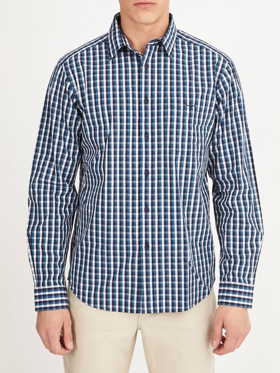 A man wears the RM Williams Collins Shirt, a 100% cotton long-sleeved blue, white, and black plaid shirt, paired with light-colored pants and standing against a plain white background.