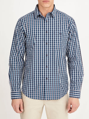 A man wears the RM Williams Collins Shirt, a 100% cotton long-sleeved blue, white, and black plaid shirt, paired with light-colored pants and standing against a plain white background.