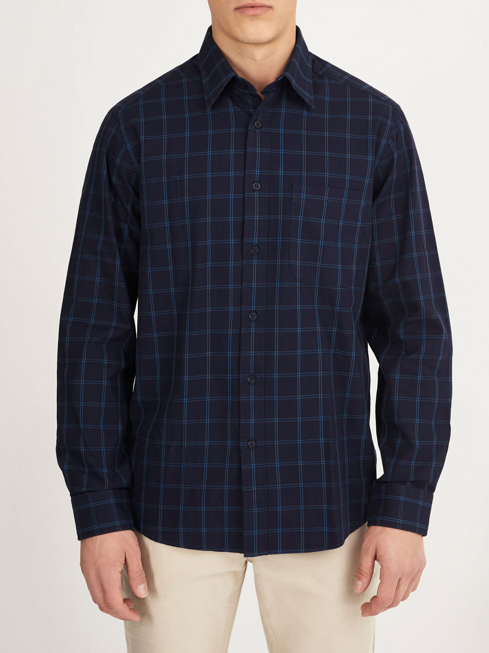 A person wearing the RM Williams Collins Shirt—a long-sleeved, navy blue checkered 100% cotton shirt with a front pocket—buttoned up with light-colored pants, stands against a plain light background.