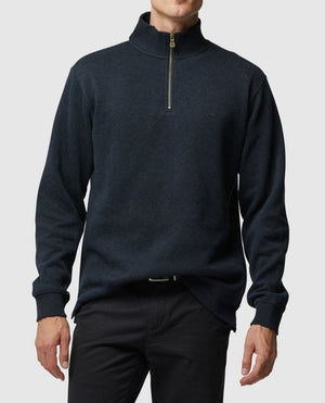 The man is dressed in a Rodd & Gunn Alton Ave Sweat, a dark half-zip design with a high collar. This stylish piece is elegantly matched with dark pants against a plain gray background.