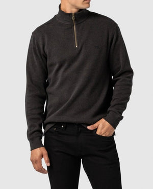 A person stands against a plain gray background wearing the Rodd & Gunn Alton Ave Sweat, a dark charcoal, relaxed fit, half-zip pullover sweater paired with black jeans. The left hand is in the jeans pocket, while the right arm rests at their side. The 100% cotton sweater features a subtle logo on the left chest.