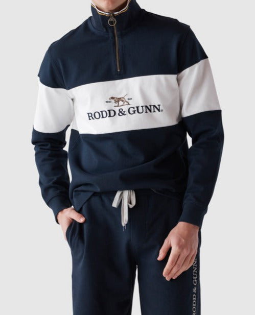 A person in a Rodd & Gunn Foresters Peak Sweat, navy blue and white half zip with "RODD & GUNN" and a dog logo on the chest, comfortably paired with matching navy blue pants, hand resting in a pocket.