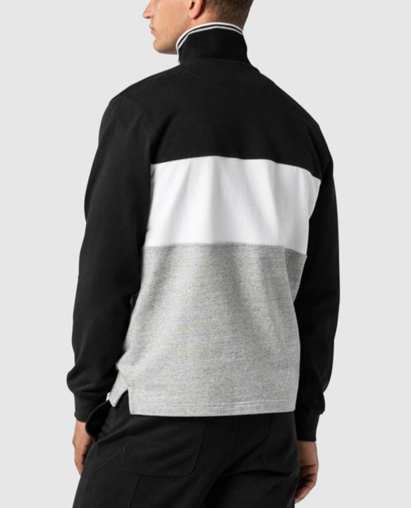 Someone wearing a Rodd & Gunn Hillop Sweat jacket with color-blocked black, white, and gray sections is shown facing away. The 100% cotton jacket includes a mock neck and half zip, paired with black pants against a plain light gray background.