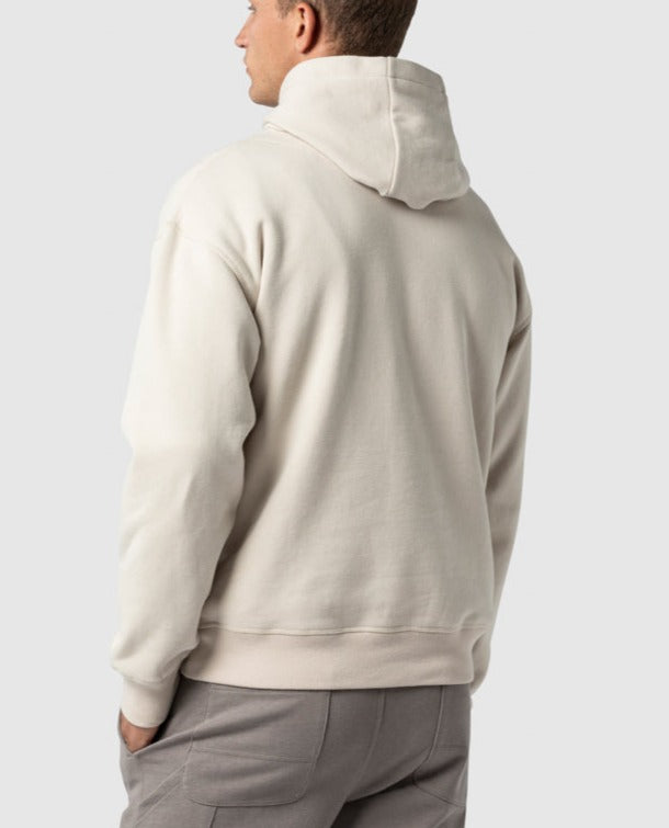 A person is seen from the back wearing the Rodd & Gunn Worser Bay Sweat cream hoodie with a soft fleece lining and light gray pants. The hood is down, their left hand by their side, against a plain light gray background that enhances the serene and understated elegance.