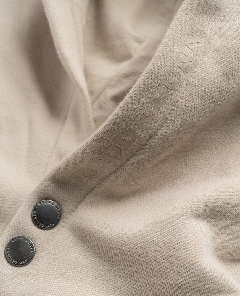 Close-up of the Rodd & Gunn Worser Bay Sweat, a beige hoodie with two engraved "Rodd & Gunn" buttons, a subtle embossed logo on soft, slightly textured fabric, and a cozy fleece lining.