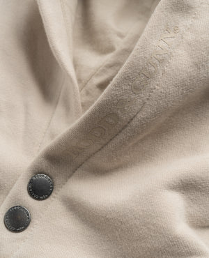 Close-up of the Rodd & Gunn Worser Bay Sweat, a beige hoodie with two engraved "Rodd & Gunn" buttons, a subtle embossed logo on soft, slightly textured fabric, and a cozy fleece lining.