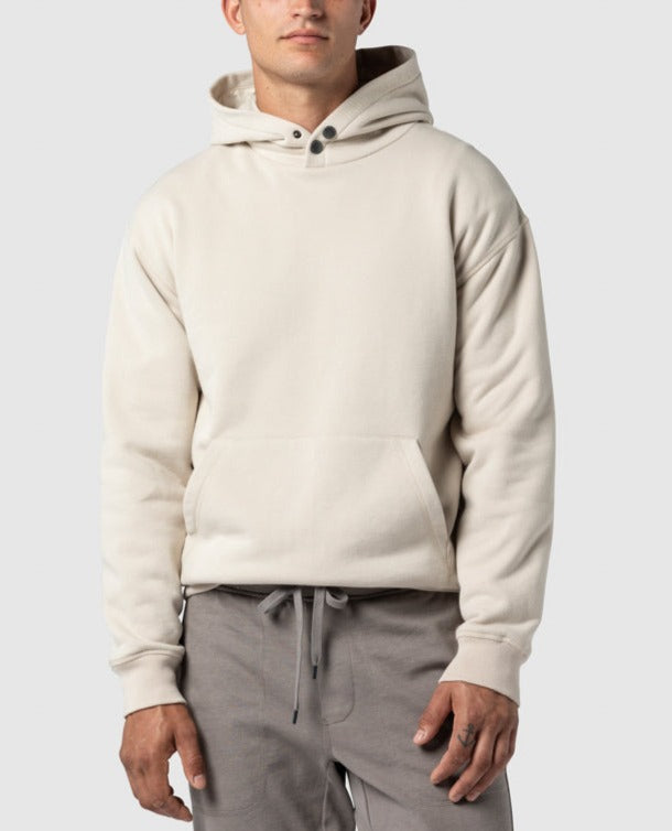A man stands confidently against a plain light gray background, embodying casual elegance in the Rodd & Gunn Worser Bay Sweat—a cream-colored hoodie with a soft fleece lining, front pocket, two button details at the collar—paired with gray drawstring pants.