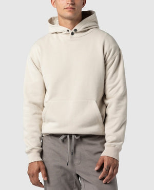 A man stands confidently against a plain light gray background, embodying casual elegance in the Rodd & Gunn Worser Bay Sweat—a cream-colored hoodie with a soft fleece lining, front pocket, two button details at the collar—paired with gray drawstring pants.