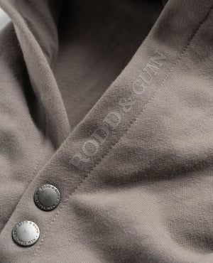 Close-up of the Rodd & Gunn Worser Bay Sweat, a light brown, comfortable hoodie with silver buttons and "FOOD & GUN" embroidered along the seam in a matching color. The high-quality soft fleece lining fabric ensures comfort. The buttons, engraved with "FOOD & GUN" around their edges, add a touch of subtle elegance to this piece.