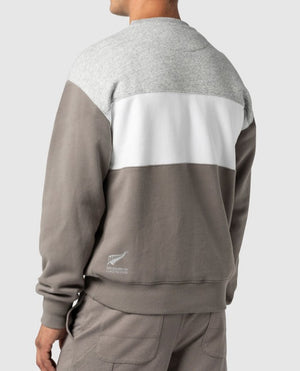 A person is wearing the Rodd & Gunn Mount Wesley Sweat, a casual color-blocked sweatshirt in gray, white, and light brown. Viewed from the back, it’s paired with light brown pants. The New Zealand national rugby team logo is on the lower left, and it features a cozy fleece lining for comfort.