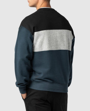 A person is seen from the back, wearing a color-blocked Rodd & Gunn Mount Wesley Sweat. The comfortable jumper features three horizontal sections: black on the top, gray in the middle, and dark blue on the bottom. The person is also wearing black pants and has their left hand in their pocket.