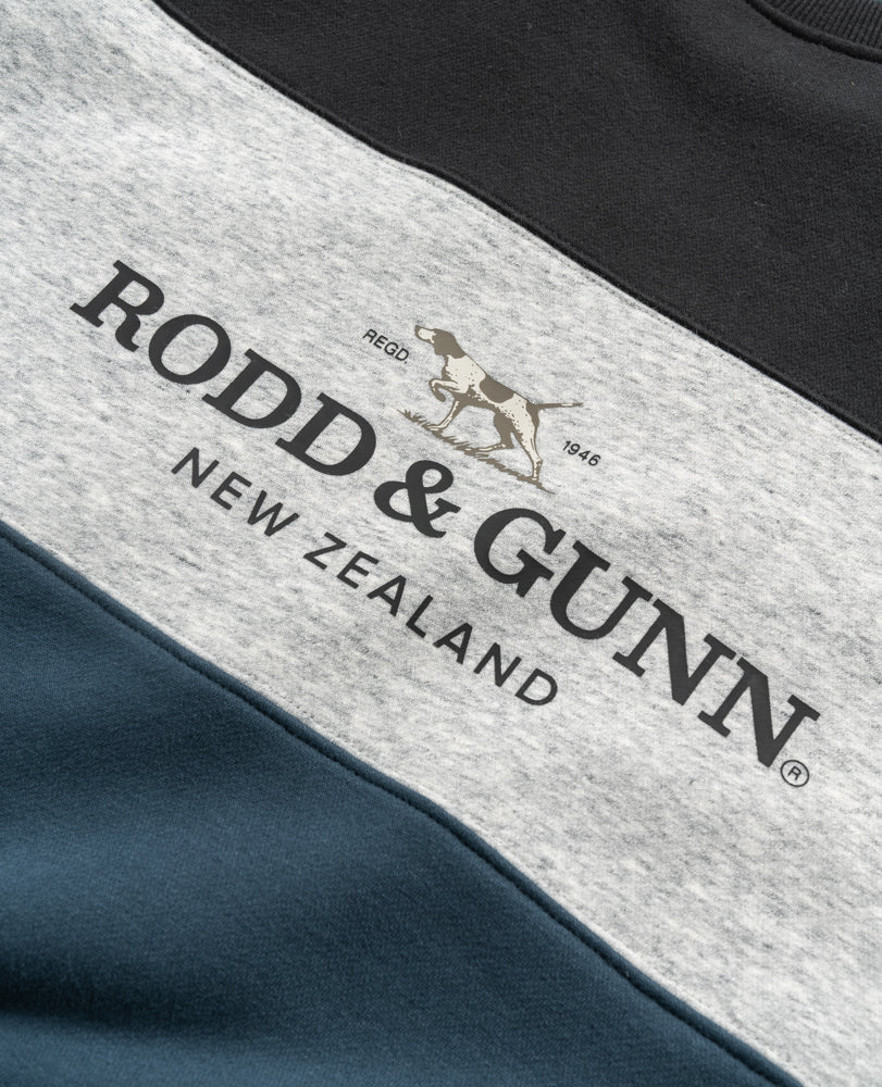 Close-up of the color-blocked Rodd & Gunn Mount Wesley Sweat jumper featuring a horizontal design with black, grey, and blue sections. The grey section showcases the brand "Rodd & Gunn New Zealand," along with a graphic of a hunting dog and the text "REGO 1946." The soft fleece lining adds extra coziness.