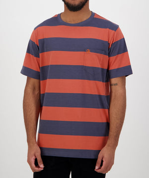 A man models a Swanndri Tomarata Stripe Tee, made from 100% cotton, with bold orange and dark blue horizontal stripes and a small chest pocket, set against a plain white background.