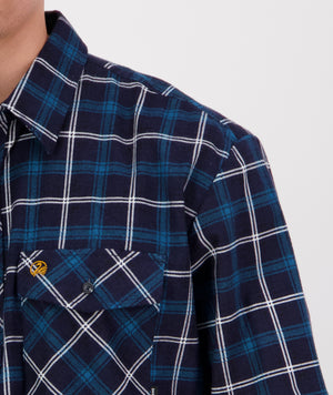 A close-up view of a person wearing a blue and white plaid long sleeve Swanndri Egmont Flannelette shirt made of brushed cotton. The shirt features a collar and a pocket with a subtle logo over the chest. Only the upper half body and part of the chest area are visible against a plain background.