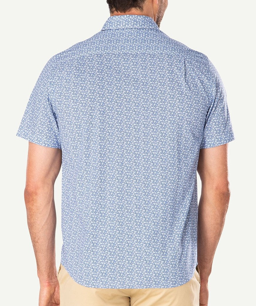 A person wearing the Gazman Casual Turtle Print Shirt, a light blue short-sleeved top with a subtle white turtle pattern. The 100% cotton, comfortable shirt features a collared neck and is tucked into beige trousers. The individual is facing away from the camera, showcasing the back of the regular fit shirt.
