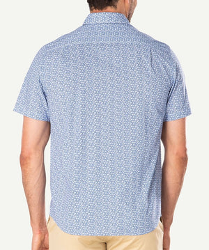 A person wearing the Gazman Casual Turtle Print Shirt, a light blue short-sleeved top with a subtle white turtle pattern. The 100% cotton, comfortable shirt features a collared neck and is tucked into beige trousers. The individual is facing away from the camera, showcasing the back of the regular fit shirt.