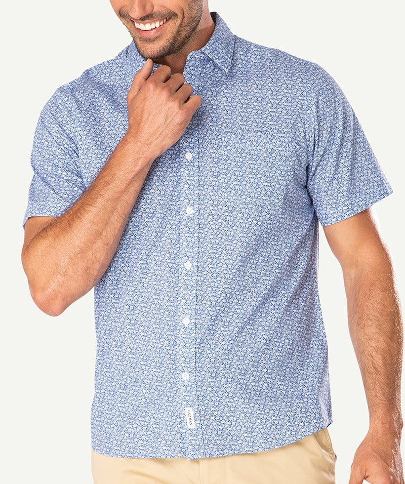 A man is wearing the Gazman Casual Turtle Print Shirt, a 100% cotton, light blue short-sleeved button-up shirt adorned with a small white floral pattern. He has light brown skin and is smiling, with his right hand touching the collar. He pairs it with regular fit light beige pants and stands against a plain white background.