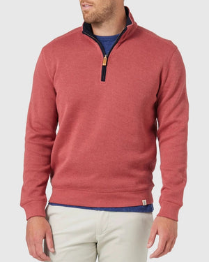 A man stands against a light gray background wearing the Blazer Heather Half Zip Sweat in red over a blue shirt and beige pants, his head slightly turned and hands relaxed at his sides.