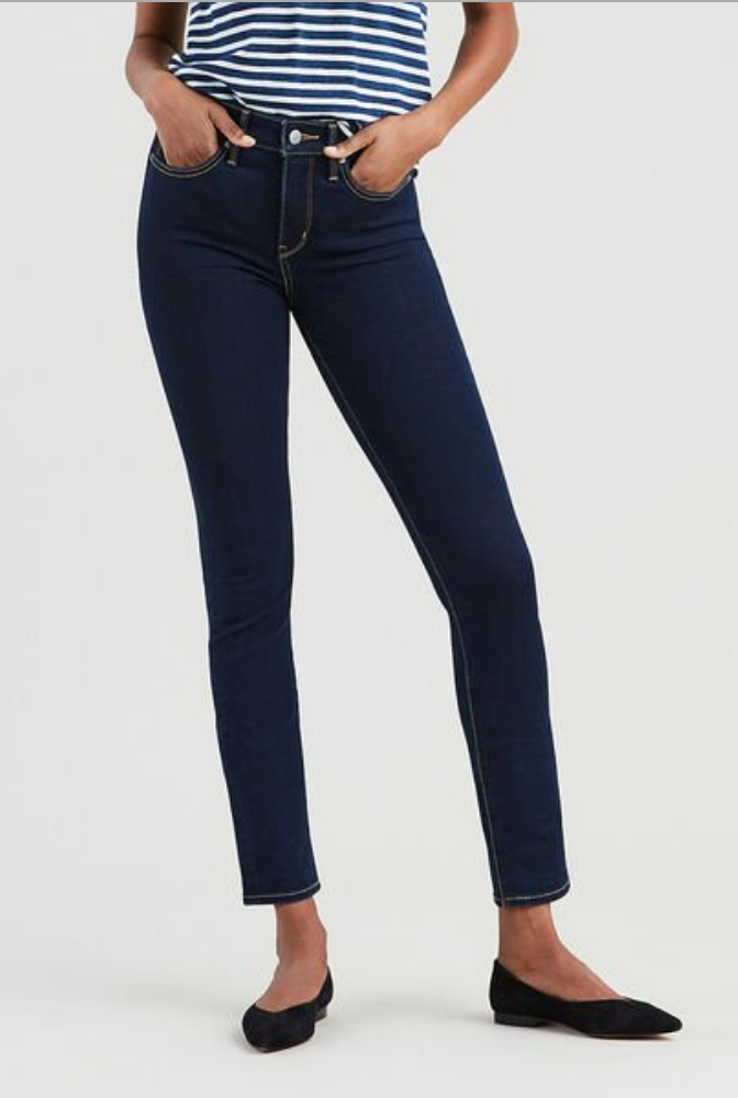 A person stands with hands in pockets, wearing dark blue Levi's 312 Shaping Slim Jean featuring a tummy-slimming panel and black ballet flats. They also sport a white and navy blue striped top. The background is plain and light-colored.