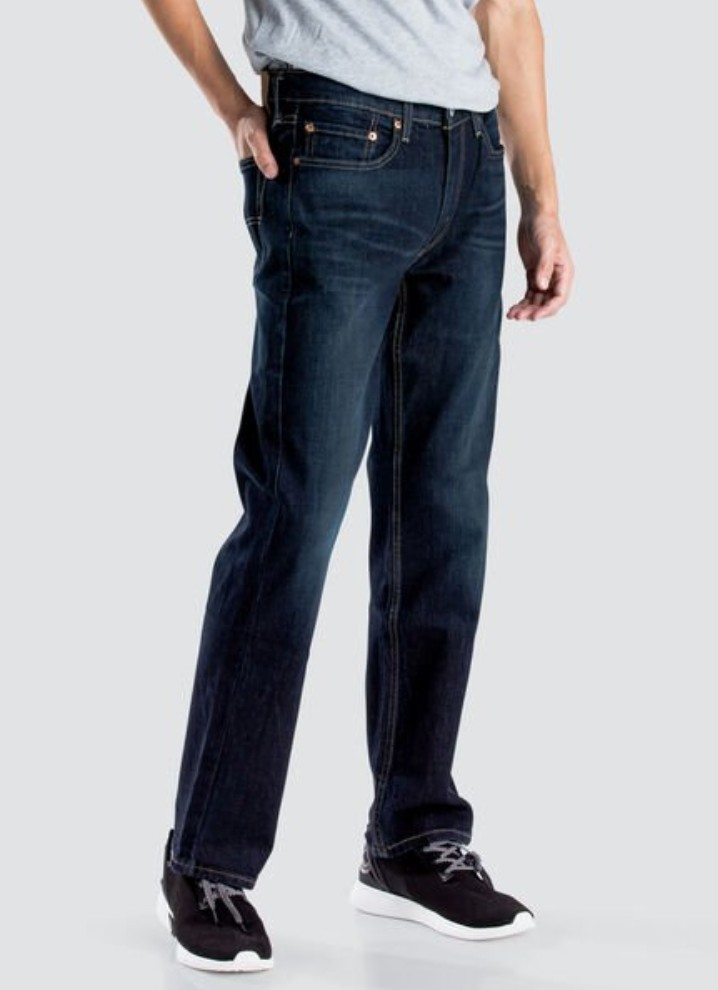 A person wearing Levis 514 Straight Jean in dark blue and a light gray shirt, with one hand in their pocket. They are also wearing black and white sneakers. The background is plain and gray.
