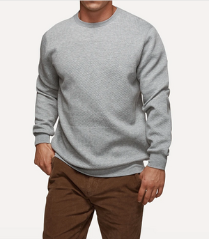 The individual is dressed in the Industrie The Robinson Crew, a light gray, long-sleeve casual jumper with a textured diagonal pattern, along with brown pants. They are situated against a plain, light background and their head is not visible in the image.