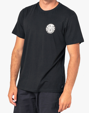A person wearing the Rip Curl Wetsuit Icon Tee, featuring a plain black design with a small Rip Curl print on the left chest, is standing against a light background. The individual has light skin, and their right arm is partially visible along their side.