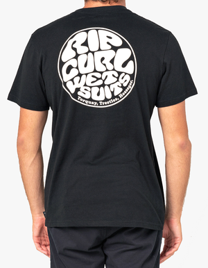 Rear view of a person wearing the Rip Curl Wetsuit Icon Tee, a comfortable black T-shirt made of 100% cotton, featuring a white, circular, retro-style Rip Curl print on the back. The logo reads "Rip Curl Wet Suit, Torquay, Trestles, Hossegor." The person is also wearing black pants.