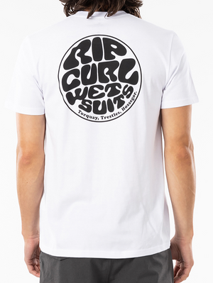 Someone with long hair is wearing the cozy Rip Curl Wetsuit Icon Tee, a white shirt made of 100% cotton that boasts a large, circular Rip Curl print on the back with the words "RIP CURL WETSUITS" and the locations "Torquay, Trestles, Hossegor." It pairs perfectly with their dark pants.