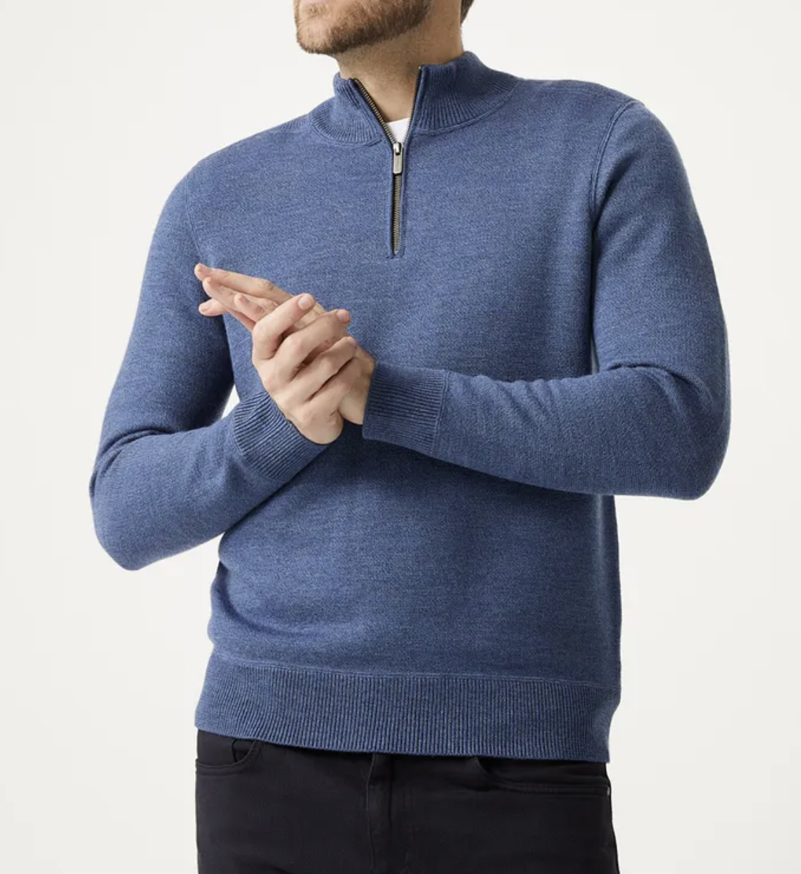 Wearing the Toorallie Grafton Half Zip Knit in blue, made from Australian Merino Wool, with long sleeves and black pants, a person stands against a plain light background with hands lightly clasped.