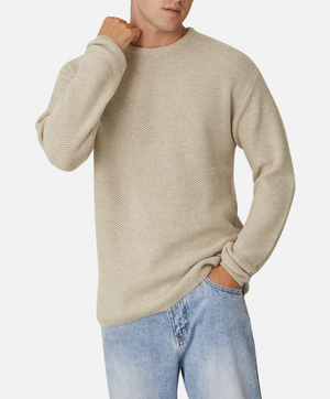 A person wearing the Industrie The Washed Culver Knit, a 100% cotton beige sweater, and light blue jeans stands against a white background. The individual has one hand in their pocket and the other adjusting the collar of the stylish knit sweater. The outfit appears casual and relaxed.