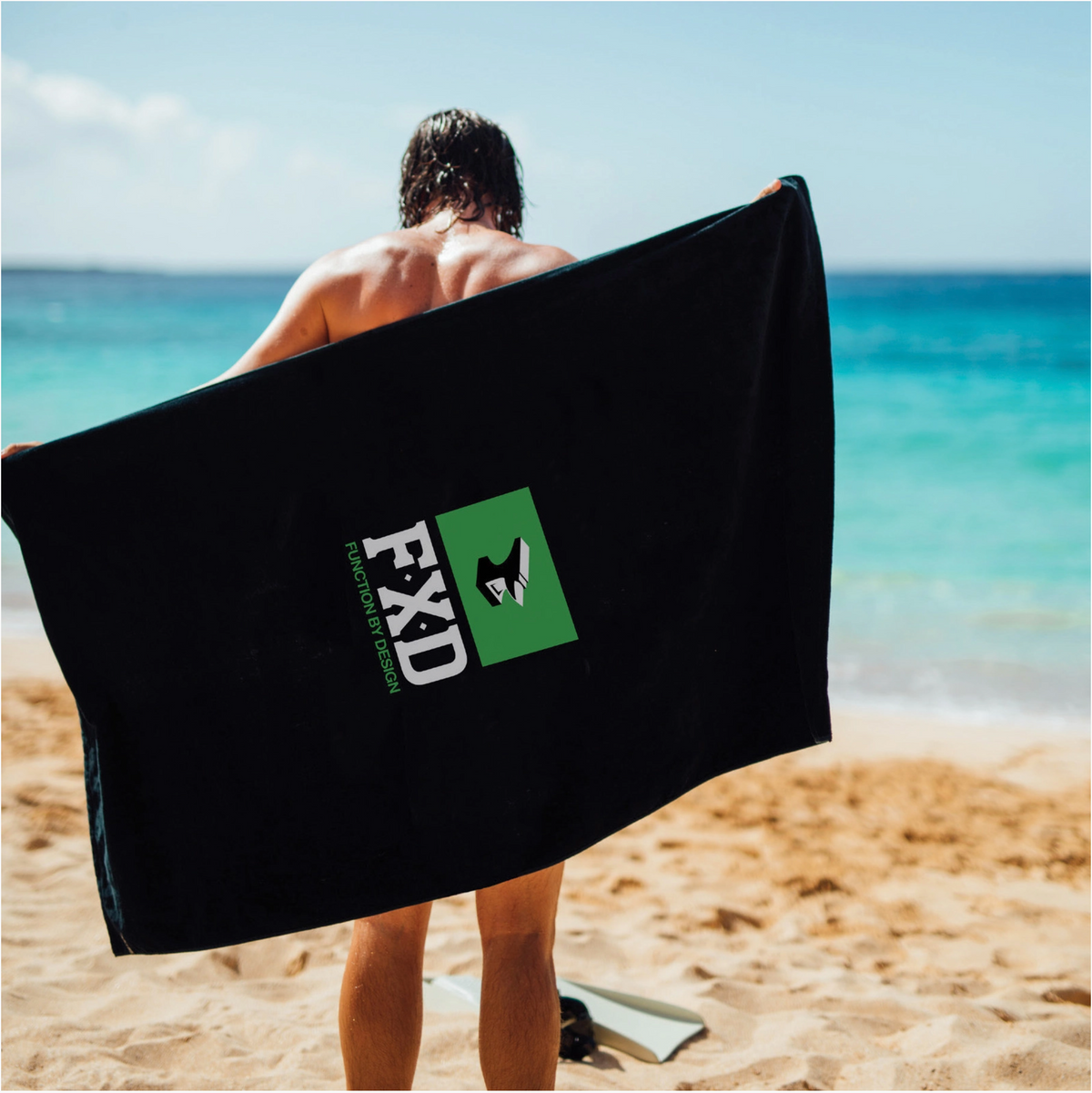 On a sandy beach with the ocean behind, someone with wet hair holds an FXD WAT-1 X Slowtide Work Towel. It's black, made from recycled polyester, and showcases a green and white logo under the clear blue sky.