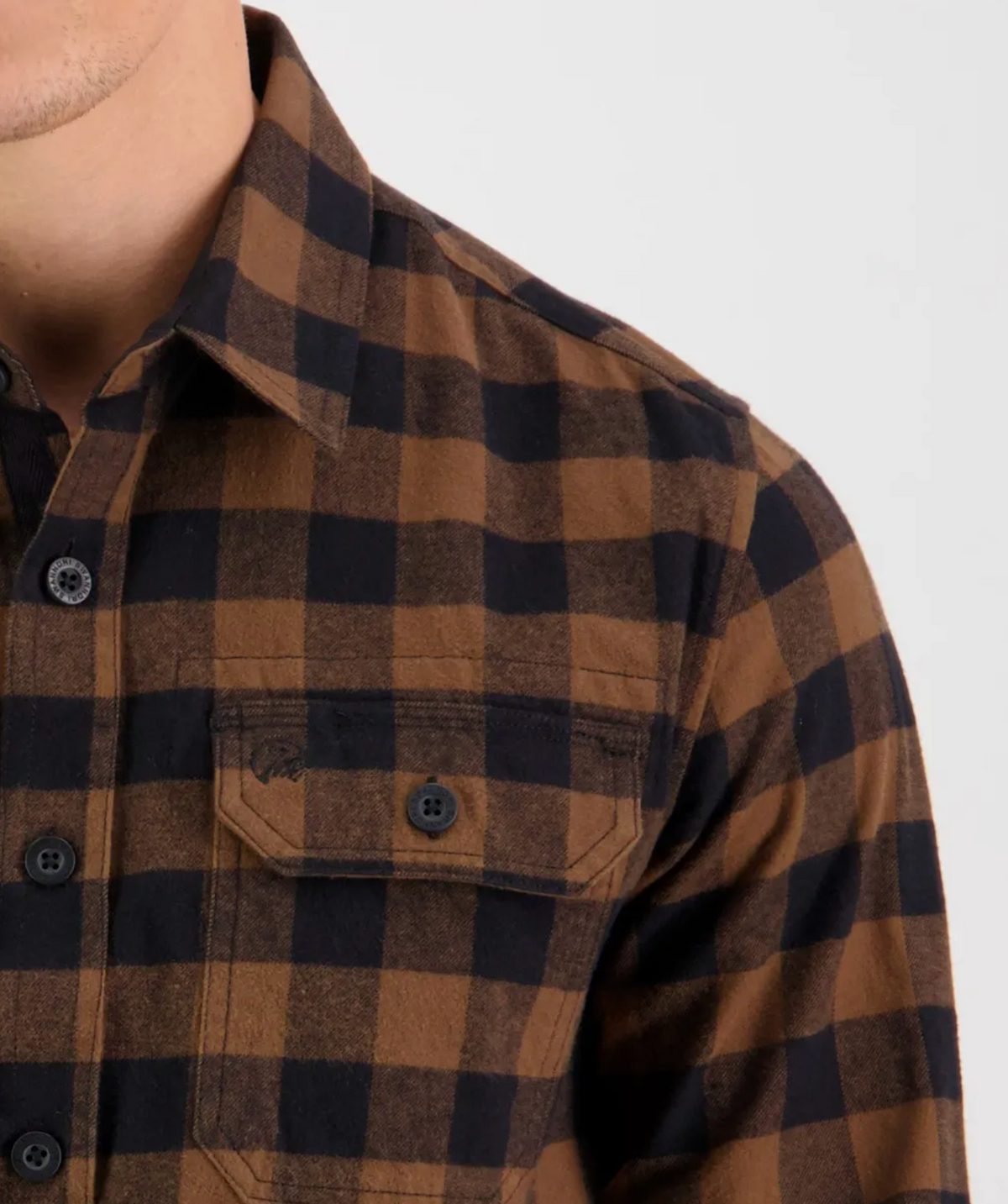 Close-up image showing part of a person wearing the comfortable Swanndri Taranaki Tailor Shirt. This brown and black checkered flannel shirt is made from 100% cotton, featuring a button-up front and a buttoned pocket on the left side of the chest. Only the lower half of the person’s face is visible at the top left corner.