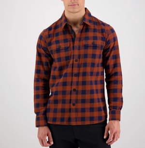 A person stands against a plain white background wearing the Swanndri Taranaki Tailor Shirt, an orange and navy blue checkered button-up flannel made from 100% cotton known for its comfort, along with black pants. The shirt features two chest pockets with button flaps. The individual's face is not fully visible.