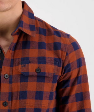 A person is wearing a Swanndri Taranaki Tailor Shirt, featuring an orange and blue checkered pattern made from 100% cotton. The focus is on the upper portion of the torso, highlighting details of the collar, buttons, and a chest pocket on the left side of this comfortable flannel shirt. The background is plain and light-colored.