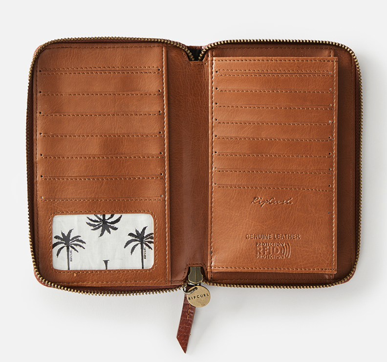 The Rip Curl Hermosa RFID Leather Wallet features a palm tree design on an open brown leather exterior, with multiple card slots, a transparent ID pocket, and a zipper around the edge. "Genuine leather protection" is embossed inside for both style and RFID protection.