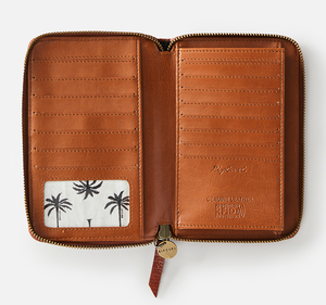 The Rip Curl Hermosa RFID Leather Wallet features a palm tree design on an open brown leather exterior, with multiple card slots, a transparent ID pocket, and a zipper around the edge. "Genuine leather protection" is embossed inside for both style and RFID protection.
