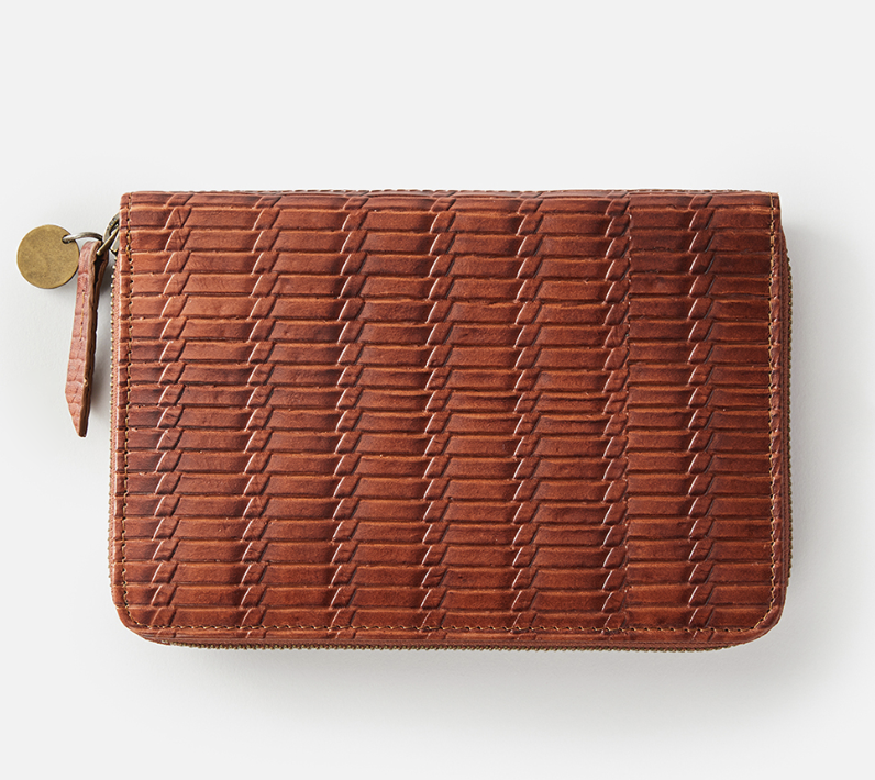 The Rip Curl Hermosa RFID Leather Wallet is brown with a textured woven pattern and zip closure. It features a small round metal tag on the zipper pull and is stylishly presented against a plain white background, offering excellent RFID protection.