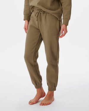 A person stands barefoot on a plain white background, wearing Rip Curl Premium Surf Trackpants in olive green. These joggers feature a cuffed design, a drawstring elastic waist, elastic cuffs, and subtle branding on the upper thigh.