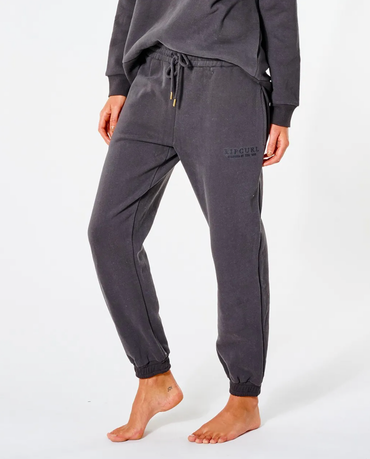 A person wears a dark, long-sleeved top and Rip Curl Premium Surf Trackpant with an elastic waist and visible logo on the upper leg. They are barefoot on a white background.