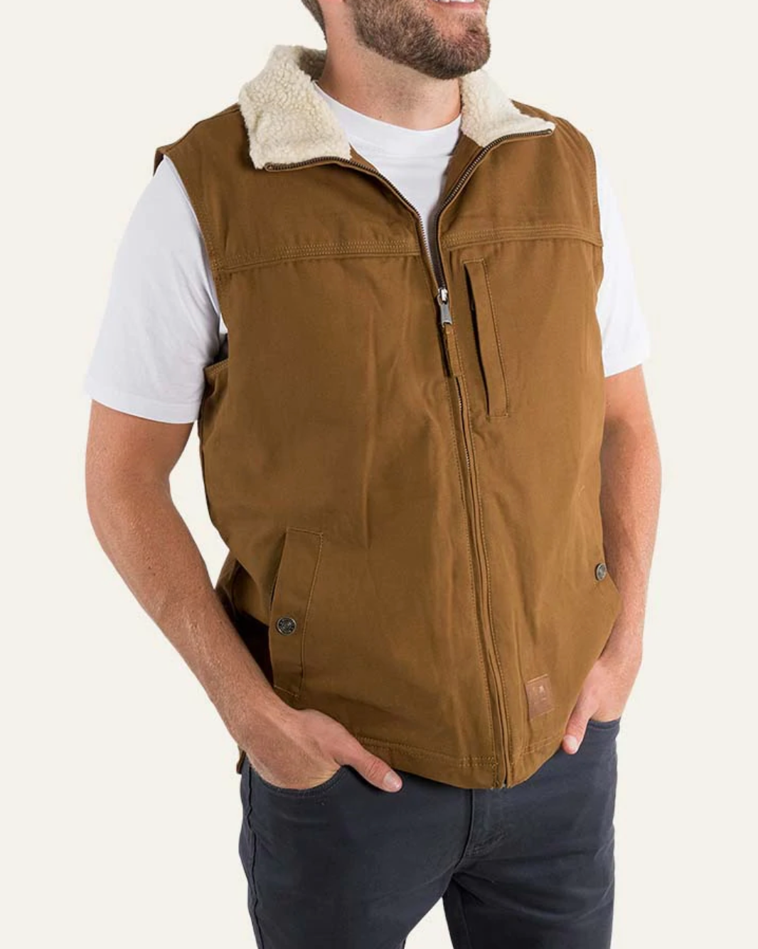 A person in a white t-shirt and dark pants wears the Ringers Western Macquarie Canvas Vest, a brown sleeveless jacket with a zippered front and two side pockets. The comfortable vest features a sherpa-lined collar. The person stands against a plain background with their hands in their pants pockets.
