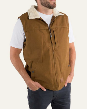 A person in a white t-shirt and dark pants wears the Ringers Western Macquarie Canvas Vest, a brown sleeveless jacket with a zippered front and two side pockets. The comfortable vest features a sherpa-lined collar. The person stands against a plain background with their hands in their pants pockets.