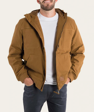 A man in a brown Ringers Western Kalgoorlie Long Canvas Jacket, white shirt, and dark pants stands smiling with hands in his jacket's multiple pockets. The unzipped jacket provides a stylish look against a plain background.