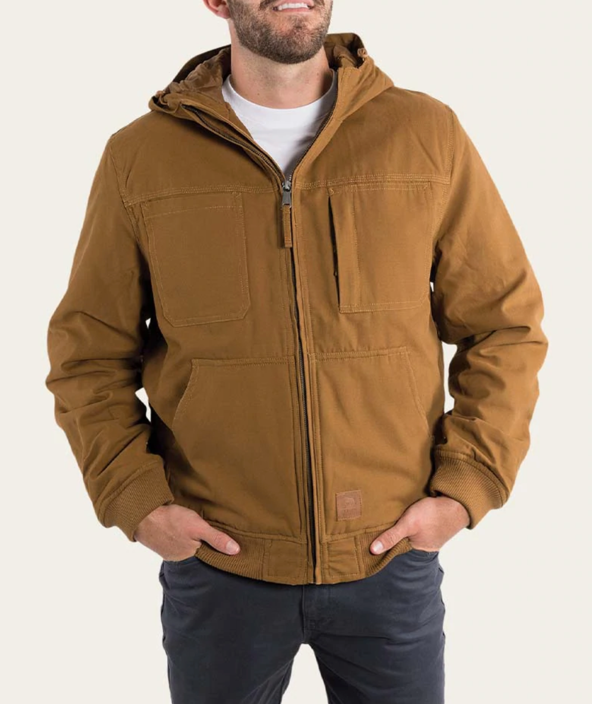 A person in a brown Ringers Western Kalgoorlie Long Canvas Jacket zipped over a white T-shirt stands against a plain background. The jacket, featuring large front pockets, is complemented by dark pants as hands are tucked into the pockets.