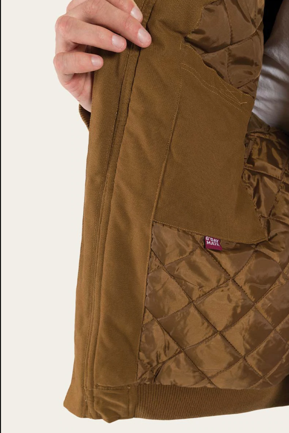 Someone holds open the brown quilted Ringers Western Kalgoorlie Long Canvas Jacket, showing its inner lining and a small red tag. The jacket, featuring diamond-pattern stitching, appears to be held by the person's left hand.