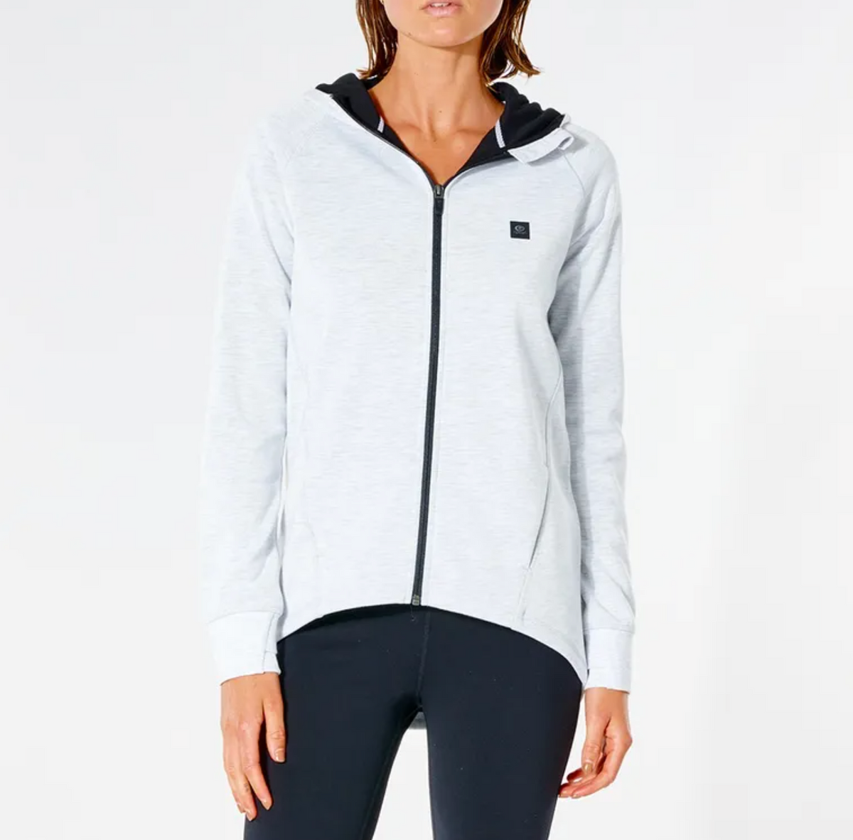 A person in a Rip Curl Anti Series Flux II Zip hoodie, light gray with a black hood lining and small chest logo, paired with black leggings against a white background, is showcasing ideal winter attire renowned for its durable water-resistant features.